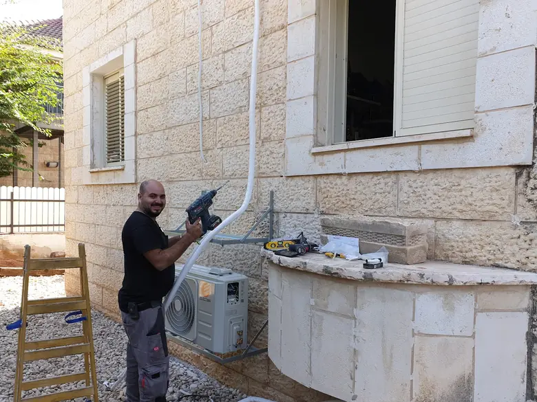 טכנאי קודח בקיר אבן ליד יחידה חיצונית של מזגן, בחצר של בית